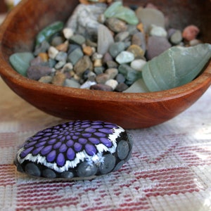 Pode incluir: Uma pedra pintada &agrave; m&atilde;o com um desenho de mandala roxo e branco. A pedra est&aacute; sobre uma toalha de mesa quadriculada branca e vermelha.