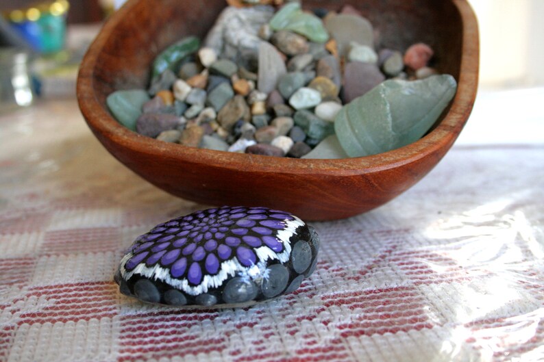 Pode incluir: Uma pedra pintada &agrave; m&atilde;o com um desenho de mandala roxo e branco. A pedra est&aacute; sobre uma toalha de mesa quadriculada branca e vermelha.