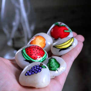 Puede incluir: Una mano sosteniendo seis piedras pintadas, cada una representando una fruta diferente: una sand&iacute;a, una pera, un racimo de uvas, un pl&aacute;tano, una naranja y una manzana. Las piedras son blancas con detalles pintados de colores.