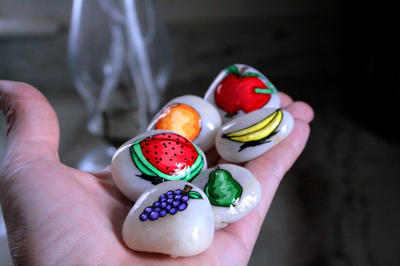 Puede incluir: Una mano sosteniendo seis piedras pintadas, cada una representando una fruta diferente: una sand&iacute;a, una pera, un racimo de uvas, un pl&aacute;tano, una naranja y una manzana. Las piedras son blancas con detalles pintados de colores.