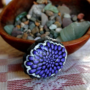 Pode incluir: Uma pedra pintada de roxo e branco com um desenho de flor. A pedra est&aacute; sobre uma superf&iacute;cie quadriculada branca e vermelha.