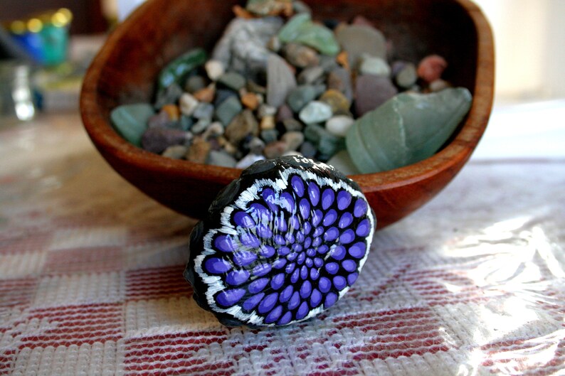Pode incluir: Uma pedra pintada de roxo e branco com um desenho de flor. A pedra est&aacute; sobre uma superf&iacute;cie quadriculada branca e vermelha.
