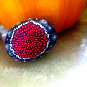 Puede incluir: Una piedra pintada a mano con fondo negro y un diseño de mandala rojo y blanco.