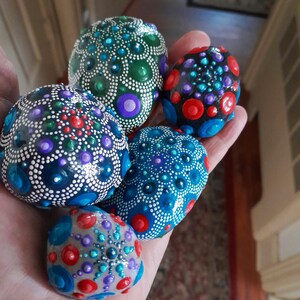 May include: Five smooth, rounded stones decorated with intricate mandala-style dot art. The stones feature vibrant patterns of red, blue, green, purple, and white paint, arranged in concentric circles and dots across their surfaces.