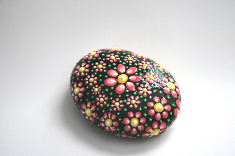 Puede incluir: Una piedra pintada con fondo negro y flores rosas y amarillas. La piedra es lisa y de forma ovalada.