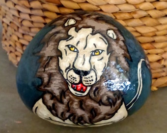 May include: Hand-painted rock art featuring a lion with a white face, brown mane, and a red tongue. The lion is painted on a blue background.
