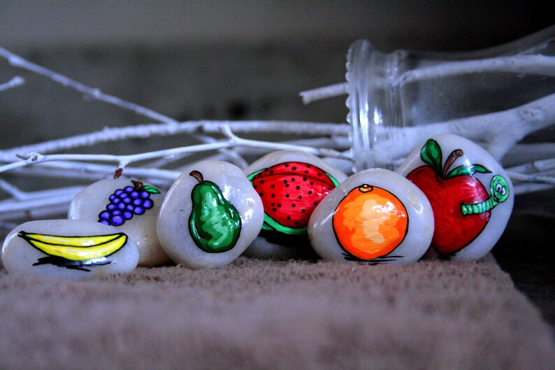 Puede incluir: Seis piedras pintadas, cada una representando una fruta diferente: un pl&aacute;tano, uvas, una pera, una sand&iacute;a, una naranja y una manzana con un gusano. Las piedras est&aacute;n dispuestas en una fila sobre una superficie marr&oacute;n.