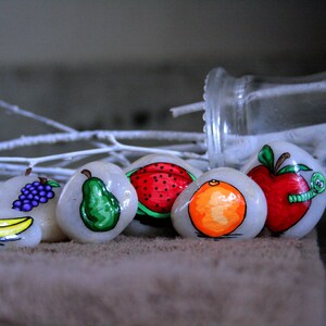Puede incluir: Seis piedras pintadas, cada una representando una fruta diferente: un pl&aacute;tano, uvas, una pera, una sand&iacute;a, una naranja y una manzana con un gusano. Las piedras est&aacute;n dispuestas en una fila sobre una superficie marr&oacute;n.