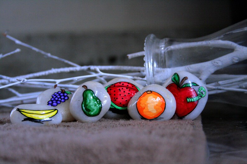 Puede incluir: Seis piedras pintadas, cada una representando una fruta diferente: un pl&aacute;tano, uvas, una pera, una sand&iacute;a, una naranja y una manzana con un gusano. Las piedras est&aacute;n dispuestas en una fila sobre una superficie marr&oacute;n.
