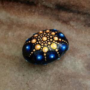 May include: A smooth, rounded stone painted with a detailed mandala pattern. The design features a dark blue base with concentric circles of gold, light yellow, and bright blue dots radiating from the center, creating a symmetrical dot art effect.