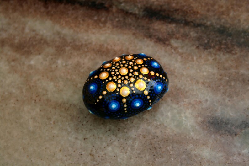 May include: A smooth, rounded stone painted with a detailed mandala pattern. The design features a dark blue base with concentric circles of gold, light yellow, and bright blue dots radiating from the center, creating a symmetrical dot art effect.