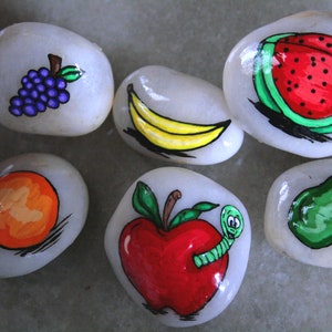 Puede incluir: Seis piedras pintadas, cada una representando una fruta diferente: un racimo de uvas moradas, un pl&aacute;tano amarillo, una sand&iacute;a roja, una naranja naranja, una manzana roja con un gusano verde y una pera verde.
