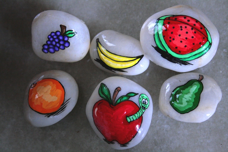 Puede incluir: Seis piedras pintadas, cada una representando una fruta diferente: un racimo de uvas moradas, un pl&aacute;tano amarillo, una sand&iacute;a roja, una naranja naranja, una manzana roja con un gusano verde y una pera verde.