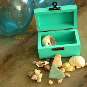 May include: An open, turquoise wooden treasure chest with a seashell inside, surrounded by various seashells and sea glass. The chest has brass hardware. A large, blue glass orb is in the background.