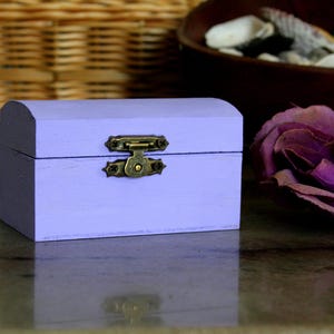 May include: A small, lavender-painted wooden chest with a curved top and a brass clasp. The chest is sitting on a reflective surface. A large, purple flower is in the background, along with a woven basket and a wooden bowl.
