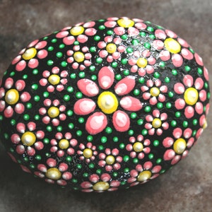 Puede incluir: Una piedra negra pintada con flores rosas y verdes. Las flores est&aacute;n hechas de puntos y tienen un centro amarillo.