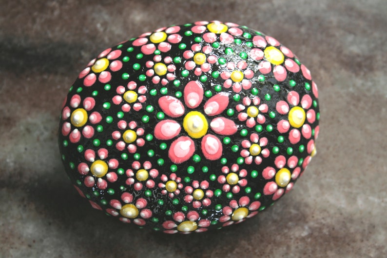Puede incluir: Una piedra negra pintada con flores rosas y verdes. Las flores est&aacute;n hechas de puntos y tienen un centro amarillo.