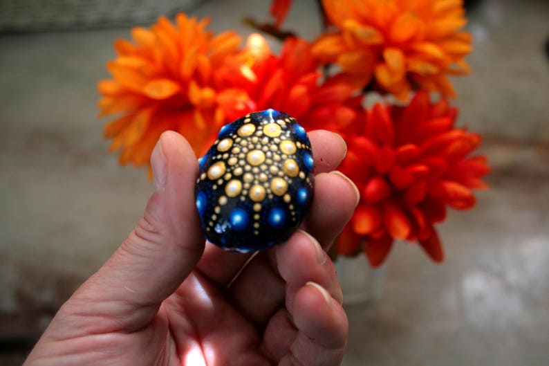 May include: A small, smooth, dark blue stone decorated with a hand-painted mandala pattern. The design features a central cluster of yellow and white dots, surrounded by concentric circles of smaller yellow dots and larger, vibrant blue dots.