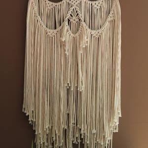 May include: A beige macrame wall hanging with a geometric design and fringe. The hanging is attached to a natural wood branch.