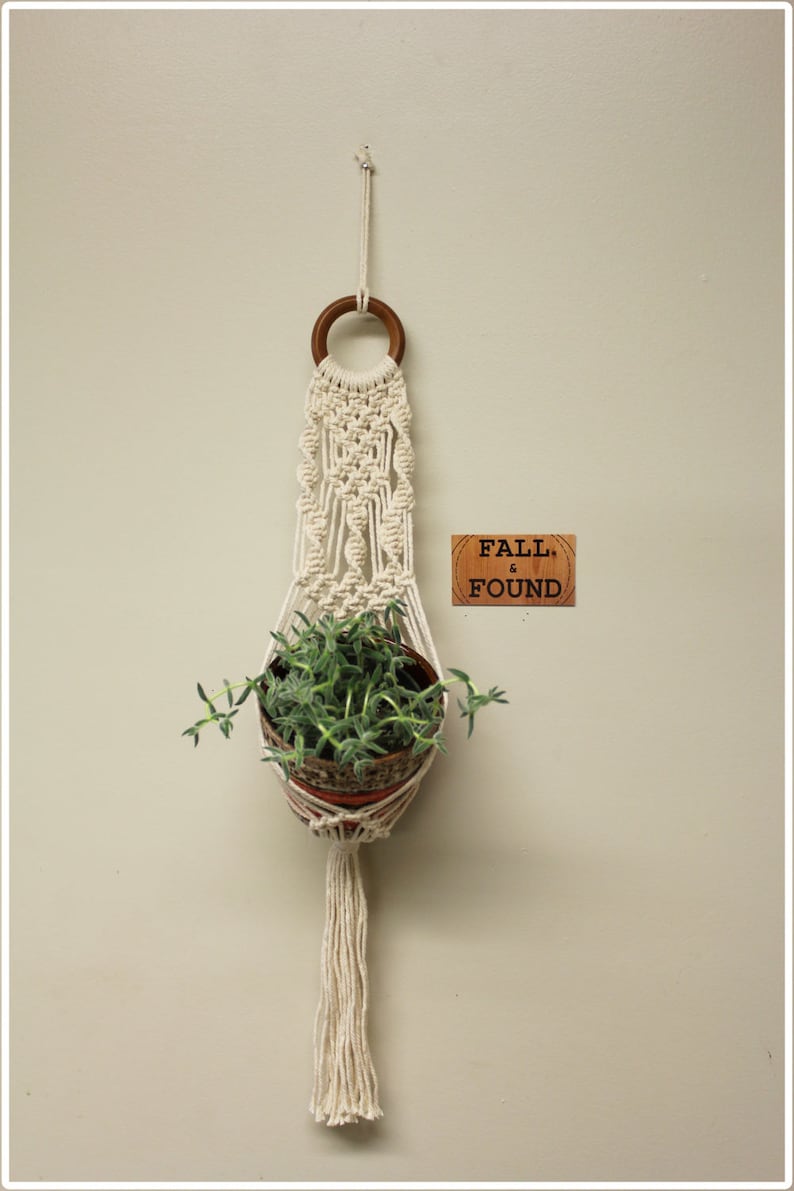Puede incluir: Un macetero de macram&eacute; blanco con un anillo de madera en la parte superior, que sostiene una peque&ntilde;a maceta de terracota con plantas verdes. El macetero tiene una borla larga en la parte inferior. Un letrero de madera con el texto "FALL & FOUND" est&aacute; fijado a la pared.