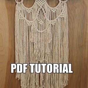 May include: A macrame wall hanging with a natural wood dowel and a cream-colored cord. The hanging features a repeating pattern of arches and fringe. The text "PDF TUTORIAL" is printed below the hanging.