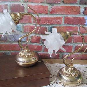 Peut inclure: Deux lampes de table vintage avec un design floral. Chaque lampe présente une base dorée et un bras incurvé, soutenant un abat-jour blanc en forme de fleur. Les lampes sont posées sur une table avec un fond en briques.