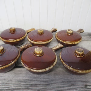 May include: Six brown ceramic soup bowls with lids. Each bowl has a handle and a lid with a knob. The bowls have a glossy finish and a contrasting beige rim. The lids have a textured knob.