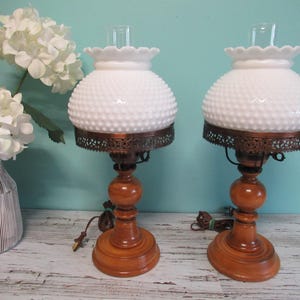 Peut inclure: Deux lampes de table vintage avec des abat-jours en verre blanc à picots et des bases en bois. Les lampes ont une bande métallique décorative autour de l'abat-jour et une cheminée en verre transparent. Les lampes sont sur une surface en bois clair.