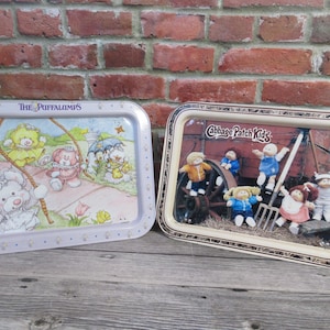 May include: Two metal serving trays with colourful illustrations. The first tray features a cartoon image of "The Puffalumps" with a purple background. The second tray features a cartoon image of "Cabbage Patch Kids" with a cream background.