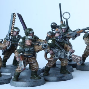 Puede incluir: Un grupo de ocho figuras en miniatura pintadas con un patrón de camuflaje, sosteniendo varias armas. Las figuras están de pie sobre bases redondas y están en una variedad de poses de acción.