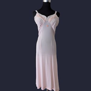 Könnte beinhalten: Ein hellrosa Vintage-Unterkleid auf einer Schaufensterpuppe. Das Kleid hat eine zarte weiße Spitzenborte am Ausschnitt und eine kleine rosa Schleife. Es hat dünne, verstellbare Träger. Das Unterkleid ist knielang und aus einem weichen, fließenden Stoff.