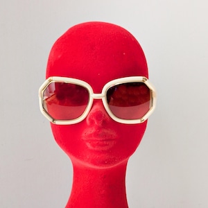 Könnte beinhalten: Cremefarbene, übergroße Sonnenbrille mit geometrischem Rahmen und dunkelroten Gläsern. Die Sonnenbrille wird auf einem roten Schaufensterkopf präsentiert, was einen auffälligen Kontrast erzeugt. Sie hat eine Retro-Vintage-Ästhetik.