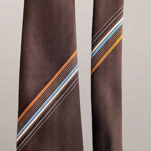 May include: A brown tie featuring diagonal stripes in orange, blue, and white. The tie is made of a textured fabric and is set against a neutral backdrop. The stripes have a dotted pattern.
