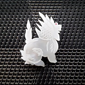 May include: A white, carved, mother-of-pearl figurine of a mythical creature with a red gemstone eye. The creature has a spiky mane and tail, and is standing on a black background.