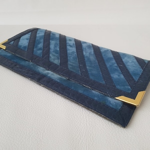 Könnte beinhalten: Eine blaue Clutch aus Wildleder mit goldenen Akzenten und einem geometrischen Muster.