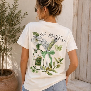 Könnte beinhalten: Ein cremefarbenes T-Shirt mit kurzen Ärmeln und einem Rückendruck, der ein Martiniglas mit Oliven, eine grüne Schleife, einen Schmetterling, eine Biene und eine kleine Olivendose zeigt. Der Text "Olive The Little Things" steht in schwarzer Schrift darüber.