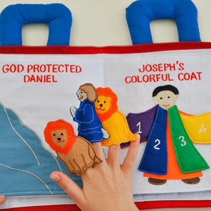 May include: A colorful felt book with two pages. The left page shows a man with a blue robe standing next to a lion. The right page shows a man with a colorful coat with numbers 1, 2, 3, and 4 on each section. The text on the left page reads "God Protected Daniel". The text on the right page reads "Joseph's Colorful Coat".