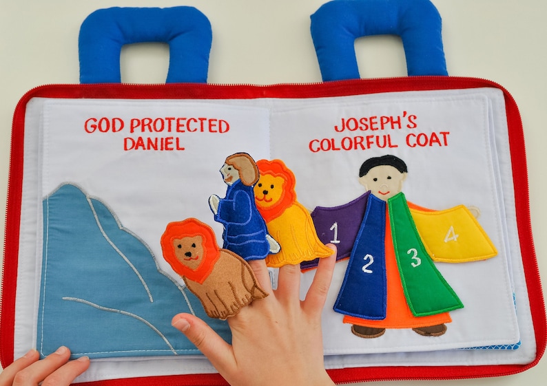 May include: A colorful felt book with two pages. The left page shows a man with a blue robe standing next to a lion. The right page shows a man with a colorful coat with numbers 1, 2, 3, and 4 on each section. The text on the left page reads "God Protected Daniel". The text on the right page reads "Joseph's Colorful Coat".