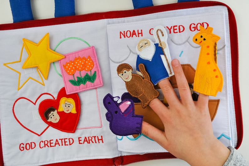 May include: A felt book for children with colorful illustrations and finger puppets. The book features a yellow star, pink tulips, a red heart with a boy and girl inside, a purple hippo finger puppet, a brown monkey finger puppet, a blue man with a white beard and a staff finger puppet, a yellow giraffe finger puppet, and the text "God created Earth" and "Noah obeyed God".