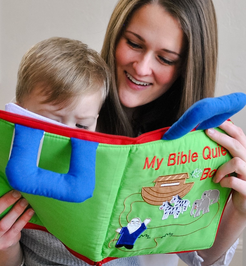 May include: A woman and a child are reading a green and blue book with the text "My Bible Quiet Book" on the cover. The book has a zipper and a handle. The book has illustrations of a boat, a zebra, and an elephant.