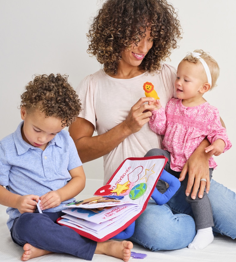 May include: A mother reading a colorful cloth book to her two children. The book has a red cover and features the text "God Created Everything".