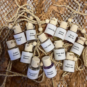 May include: An assortment of small, clear glass bottles with wooden caps, each labeled with a different fragrance name. The bottles are arranged on a textured brown surface and strung together with a decorative cord.