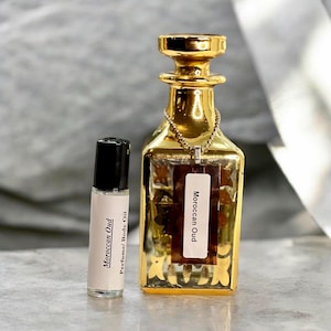 May include: A small clear glass roller bottle with a black cap and a white label reading Moroccan Oud Perfume/Body Oil next to a larger square glass bottle with a gold finish, a gold stopper, and a tag reading Moroccan Oud on a gold chain.