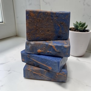 May include: A stack of four rectangular bars of soap with a deep blue base color and swirling copper-colored accents. The top bar has a textured, granular surface, while the lower bars feature smooth surfaces with visible streaks of copper-toned swirls.