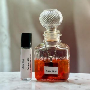 May include: A small cylindrical perfume oil roller bottle with a black cap and a white label reading Rose Oud Perfume Oil IG@agaveworldco, next to a square glass decanter with a textured stopper containing orange liquid and a Rose Oud pendant.
