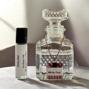 May include: A clear glass perfume oil roller bottle with a black cap and a white label reading White Oud and Perfume Oil IG@agaveworldco, next to a clear glass decanter with a diamond-cut pattern and a square pendant labeled White Oud.