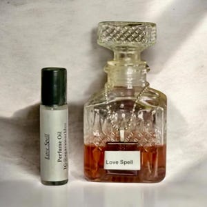 May include: A small cylindrical perfume oil roller bottle with a black cap and a label reading Love Spell Perfume Oil IG@agaveworldco next to a square glass decanter with a diamond-patterned texture, a clear stopper, and a Love Spell label.