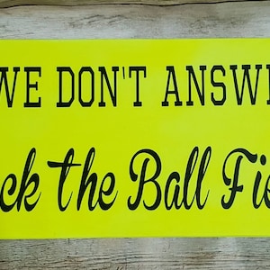 May include: Yellow wooden sign with black text that reads "If we don't answer, check the ball field!" with red and white stitching on either side of the text.