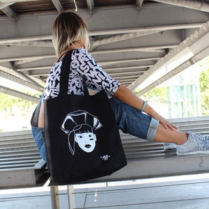 May include: A woman wearing a white and black patterned shirt and blue jeans carries a black tote bag with a white graphic of a woman's face. The bag has a white strap.