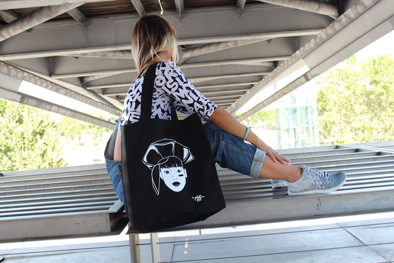 May include: A woman wearing a white and black patterned shirt and blue jeans carries a black tote bag with a white graphic of a woman's face. The bag has a white strap.